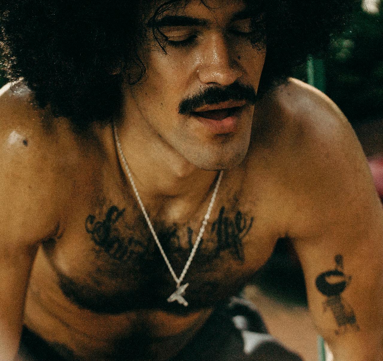 Close-up of a shirtless man with a mustache and afro, showcasing tattoos and jewelry.